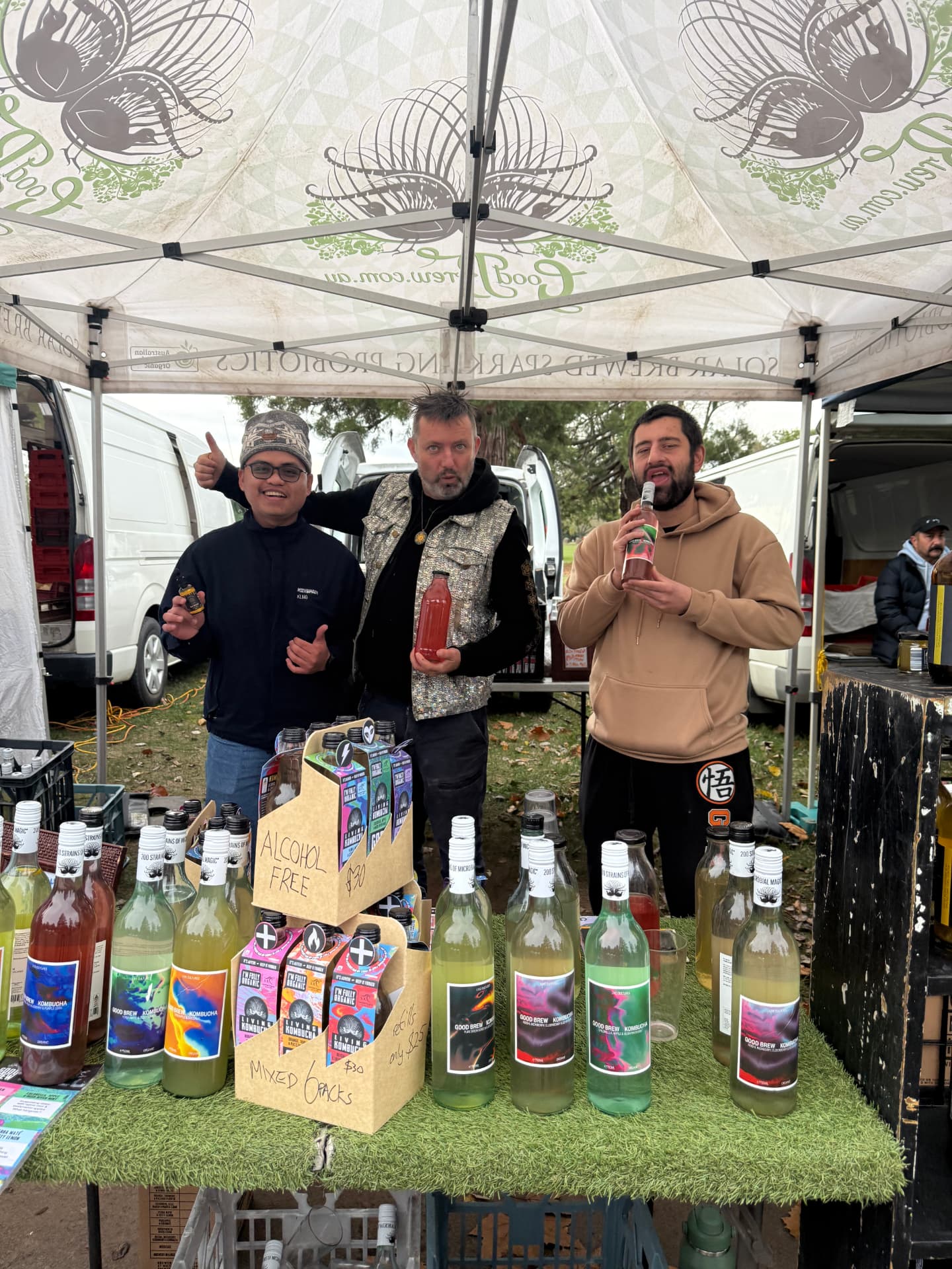 Three people smiling behind a market stall stacked with bottled drinks